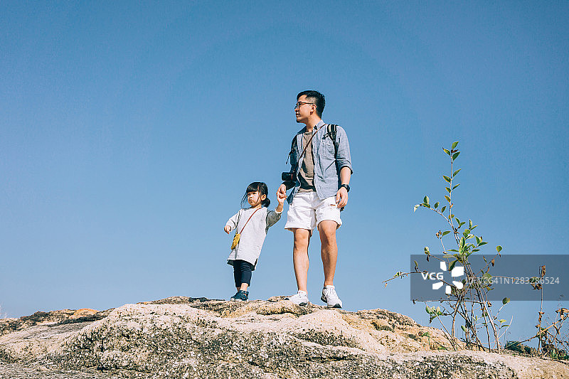年轻的亚洲父亲牵着小女儿的手爬山，站在清澈的蓝天下。他们玩得很开心，一起度过了一段高质量的亲密时光。在一个阳光明媚的日子里，享受大自然和美丽的风景图片素材