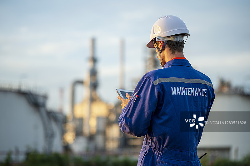 石油维护工程师与数字板在石油和天然气炼油厂设施可见的背景后视图。可持续能源的概念。图片素材