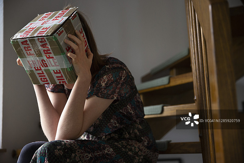 一个十几岁的女孩坐在楼梯上，头上顶着一个标着“易碎品”的纸箱图片素材