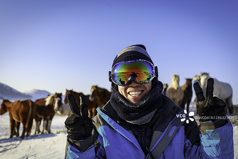 一个穿着冬季夹克，戴着滑雪眼镜的亚洲男人，在马牧场的雪地上，对着镜头开心地笑着图片素材