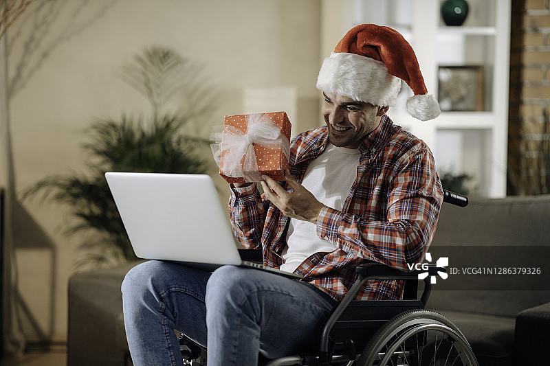 一个快乐的年轻人在轮椅上与圣诞老人的帽子在一个视频电话祝愿圣诞快乐的股票照片图片素材