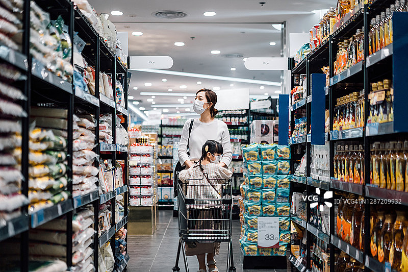 可爱的亚洲小女孩坐在购物车里和她的妈妈在超市购物。他们正沿着商品通道购物，两人都戴着防护口罩图片素材