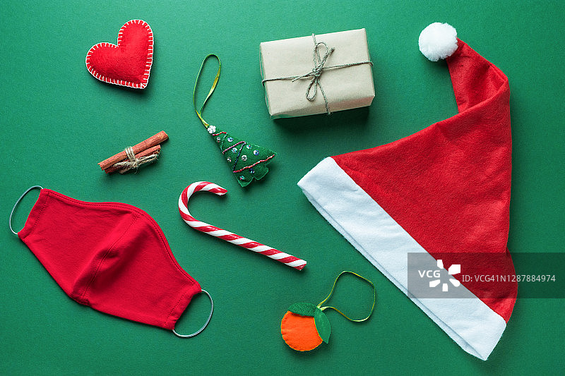 红色医用防护口罩，圣诞老人帽或帽，条纹糖果，手工圣诞树玩具，肉桂，绿色背景礼物。预防和预防COVID - 19大流行的传播。快乐健康圣诞的理念。图片素材
