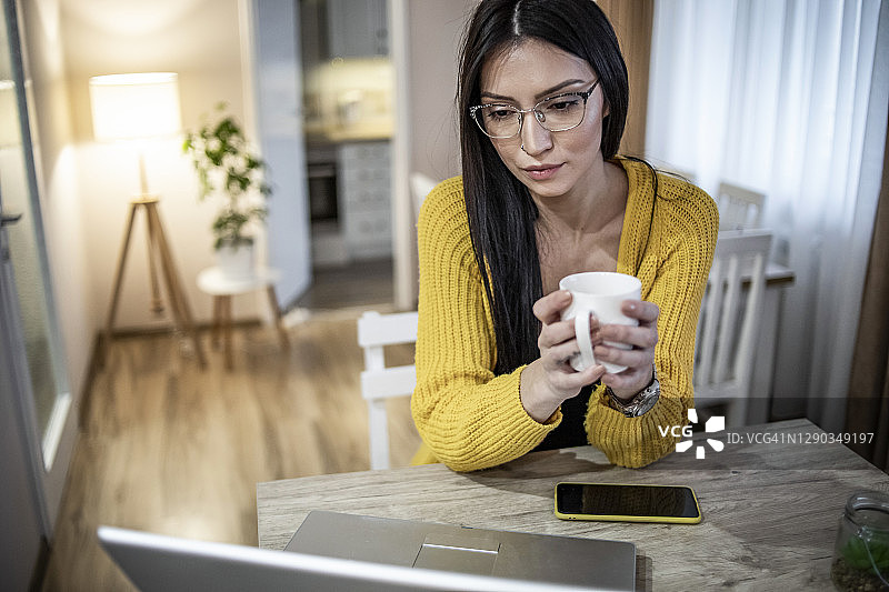 一位年轻女子在家里用一台电脑和一部智能手机工作。一位穿着漂亮黄色毛衣的黑发美女坐在家里，用智能设备在网上工作图片素材