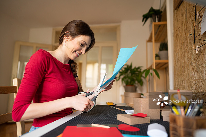 微笑的黑发女人在她的工作台上用剪刀剪纸，为礼品盒做装饰图片素材