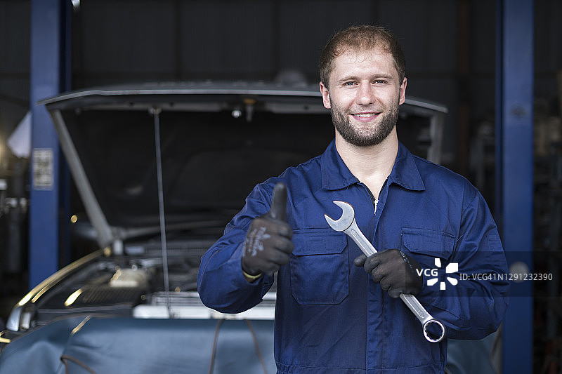 这是一幅技工在汽车修理店修理汽车的照片，他拿着扳手微笑着看着相机。图片素材