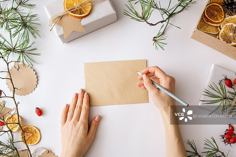 圣诞空白贺卡模型。一个女人在写一封空白的节日冬季作文。工艺信封，礼品盒，白色背景上的自然装饰。平铺，俯视图，拷贝空间。图片素材