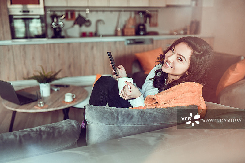 微笑的女性享受舒适的时间在家里毯子，咖啡和智能手机图片素材