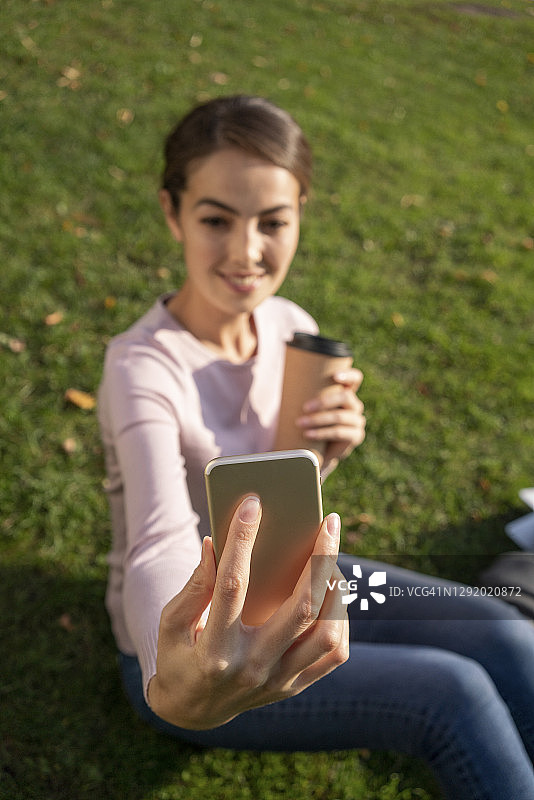 秋天，年轻女企业家坐在公园草地上用智能手机自拍图片素材