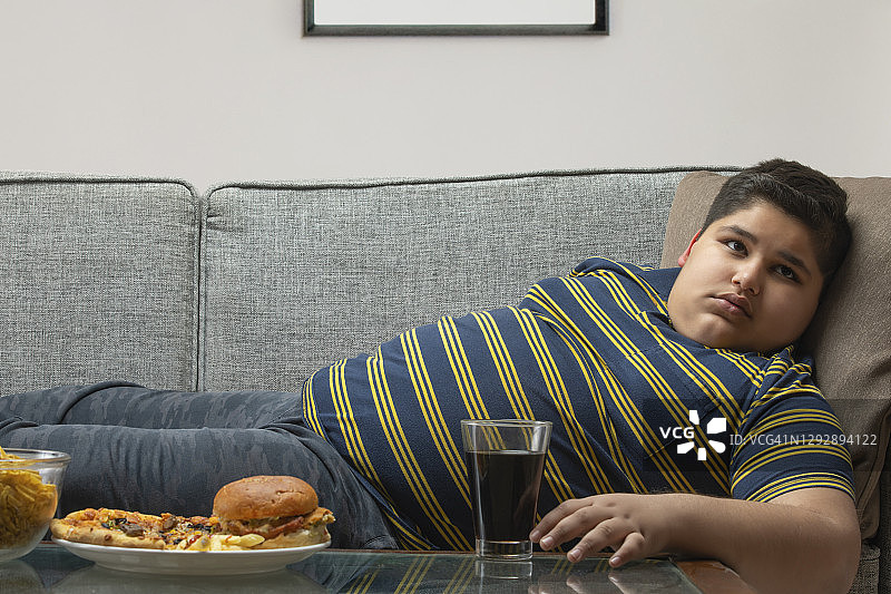 一个小男孩躺在沙发上，桌上放着垃圾食品。(肥胖)图片素材