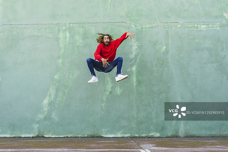 年轻男子的肖像穿着红色运动衫跳在绿色墙前的空气图片素材