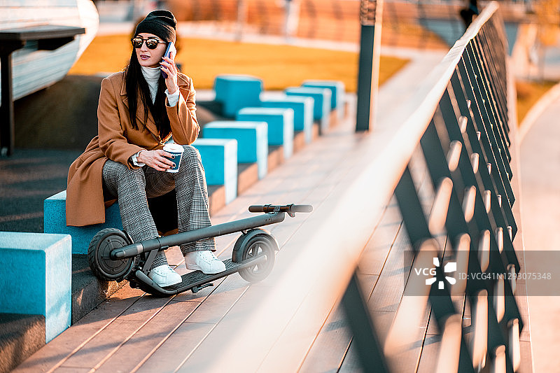 现代女商人在喝咖啡休息时坐在折叠的电动摩托车旁讲电话图片素材