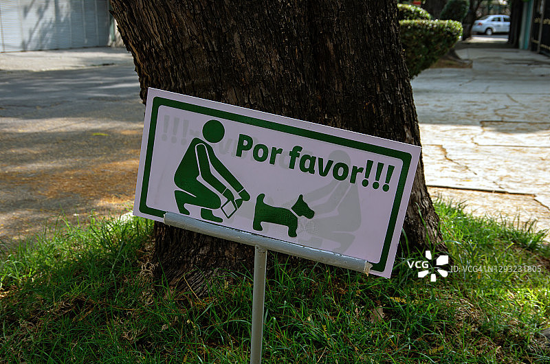 一个西班牙语的标语插在一棵街道树旁的草地上，要求宠物主人捡起自家狗的粪便图片素材