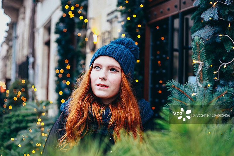 红头发的年轻快乐微笑的女人穿着温暖的冬装在大街上，圣诞节日市场装饰着仙女灯和松树。生活方式度假。图片素材