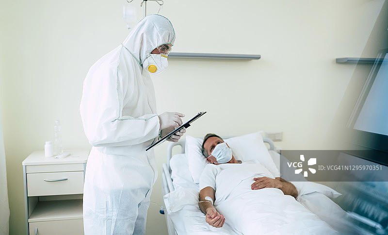 一名成年男性病人躺在医院病房的床上，由身穿防护服和戴着口罩的医生进行检查。图片素材