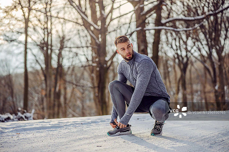 在雪天里，跑步者蹲在大自然的小路上系鞋带。健康的生活方式，运动服，下雪的天气图片素材