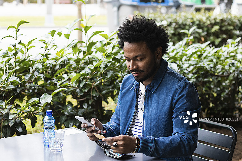 一名留着非洲式发型的男子在路边咖啡馆用信用卡和智能手机付款图片素材