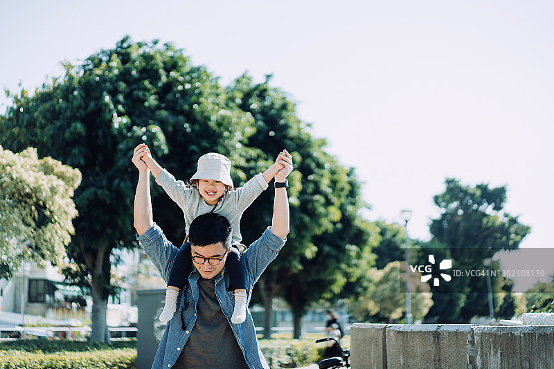 一位慈爱的年轻亚洲父亲肩上扛着小女儿在户外。小女儿开心地笑着。在一个阳光明媚的日子里，他们在一起度过了一段美好的时光。家庭生活方式图片素材