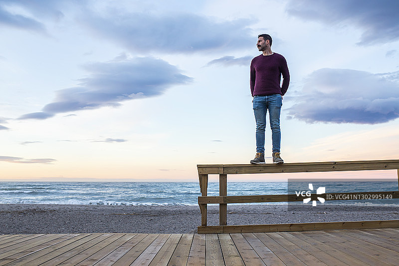 一名男子双手插在口袋里，背靠天空站在海滩的木栏杆上图片素材