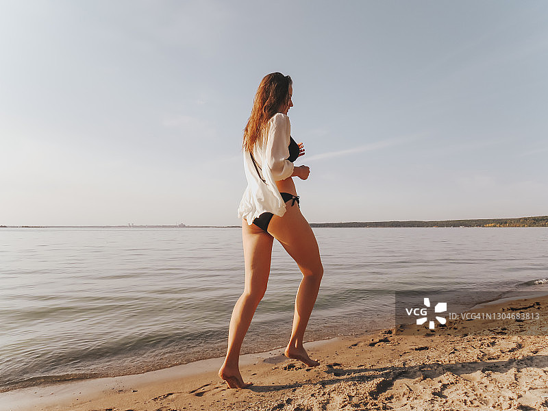 一位迷人的年轻女子穿着黑色比基尼和丝绸衬衫，晒黑了的修长腿在日落时分沿着海边沿着水边跑图片素材