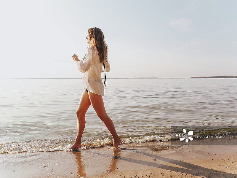 日落时分，一个迷人的年轻女子穿着黑色比基尼和丝绸衬衫，晒成古铜色，身材苗条，站在沙滩上的水边图片素材