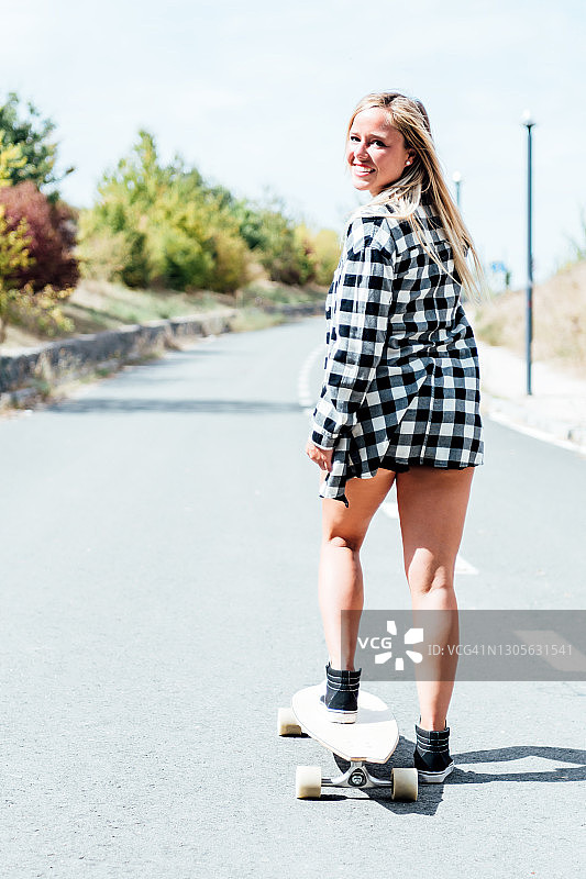 金发女人与她的背部在短裤和检查衬衫滑冰在她的长板滑板。图片素材