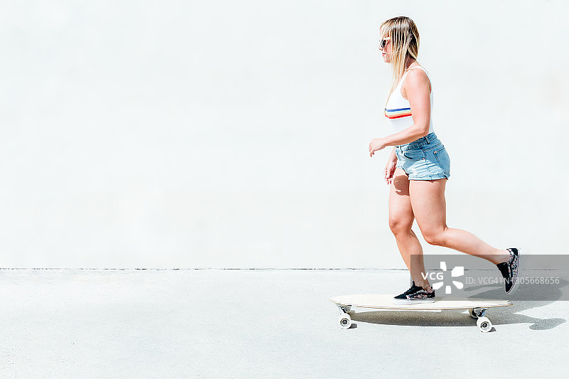 金发女人在短裤与黑色太阳镜滑冰与她的长板滑冰，白色的背景。图片素材