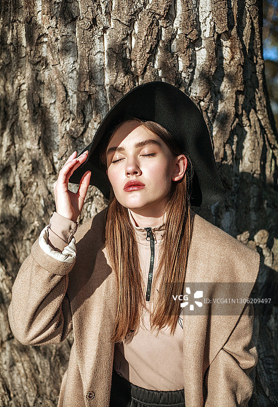 时尚时尚的年轻女士在米色外套和黑帽子在城市公园的背景树皮，美丽的女人肖像图片素材