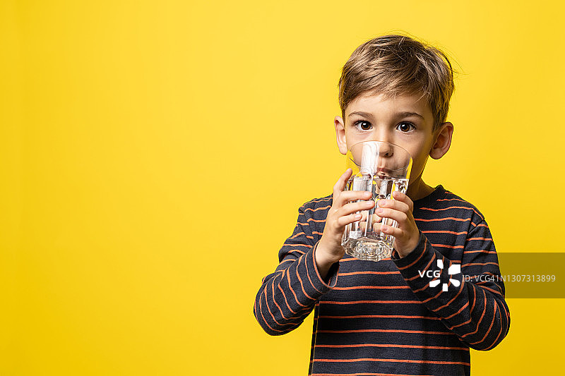 小白种男孩小男孩拿着一杯水，站在现代黄色背景前喝着水，看着有拷贝空间的相机图片素材