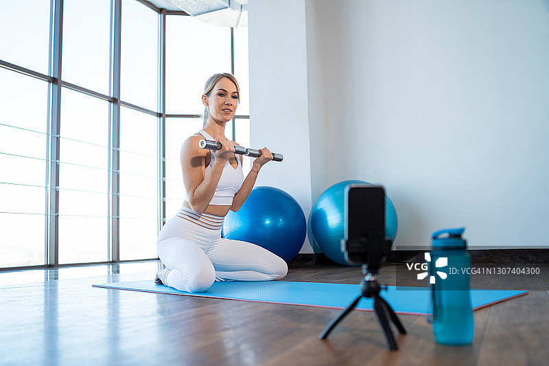 开朗的女性健身博主用智能手机摄像头上的哑铃记录锻炼图片素材