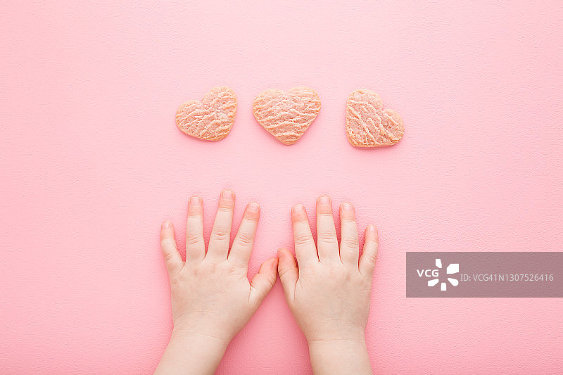 小女孩的手和心形的干饼干在浅粉色的桌子背景。柔和的颜色。甜的零食。特写镜头。视角拍摄。自顶向下的观点。图片素材