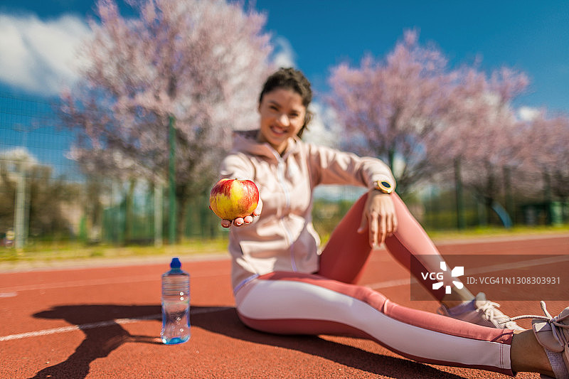 运动的年轻女子在跑道上跑步后吃苹果。图片素材