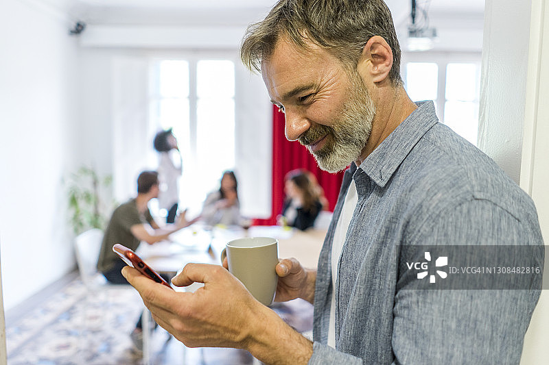 微笑的男性企业家在办公室的咖啡休息时间使用智能手机图片素材