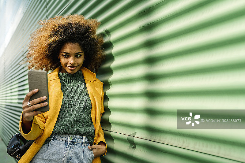 笑容满面的非洲裔女性双手插在口袋里，倚靠在绿色墙壁上用智能手机自拍图片素材