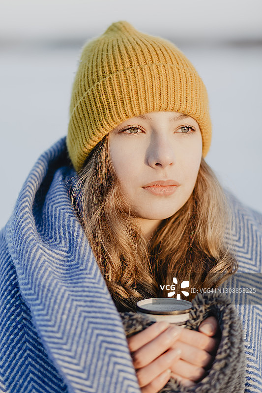 肖像美丽的少女戴着针织帽暖和自己与羊毛毯子和一杯热茶图片素材