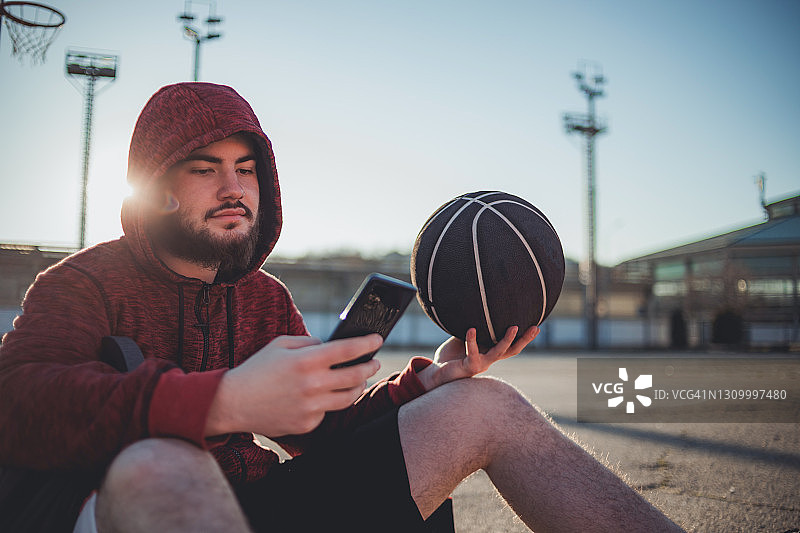 疲惫的年轻人坐在篮球场上，看着周围的环境，骄傲地等待下一次训练——这是成功的动力。图片素材