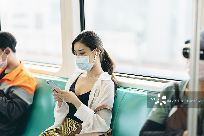 亚洲女性在乘坐地铁或火车上下班时戴口罩、使用智能手机和耳机，保持社交距离图片素材