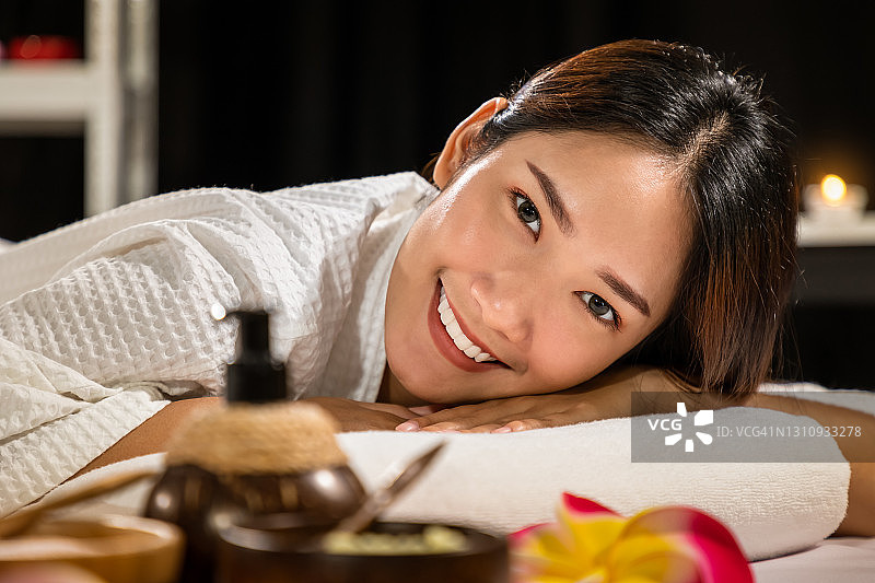 美丽年轻的亚洲女人休息在床上水疗，与按摩油，磨砂粉，和蜡烛，看着相机。图片素材
