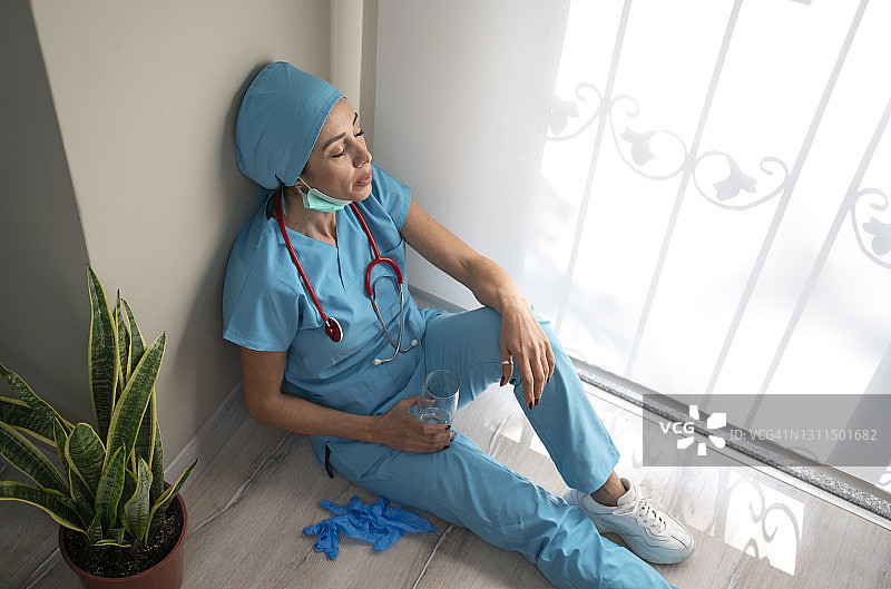 过度劳累的女医务人员戴着口罩、蓝色制服和手套坐在医院的地板上图片素材