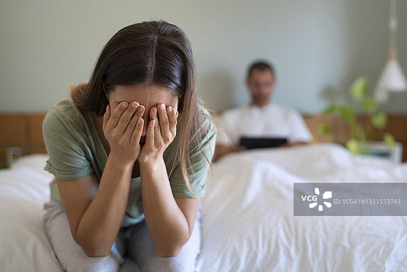 绝望的女人捂着脸坐在床上，而男朋友在家里用平板电脑做背景图片素材