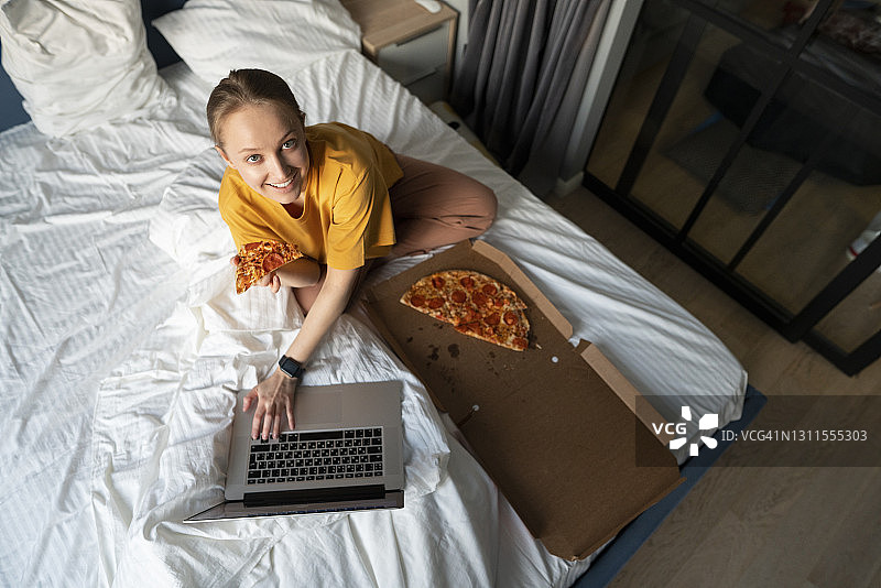 带着披萨微笑的年轻女子坐在床上的笔记本电脑旁图片素材