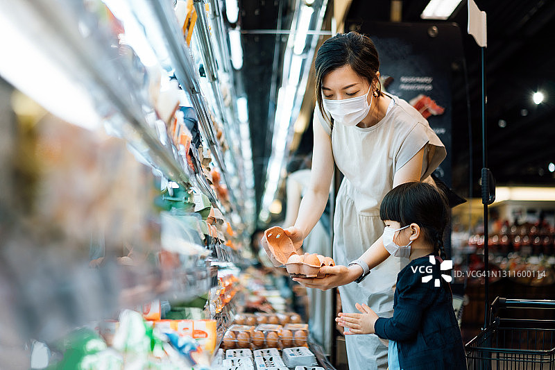 年轻的亚洲母亲和可爱的小女儿带着防护口罩一起在超市购物，在乳制品区选择新鲜的有机鸡蛋。家庭生活方式，健康饮食习惯图片素材