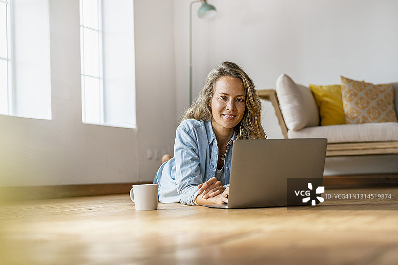 一个面带微笑的女人躺在家里的硬木地板上，在笔记本电脑前图片素材
