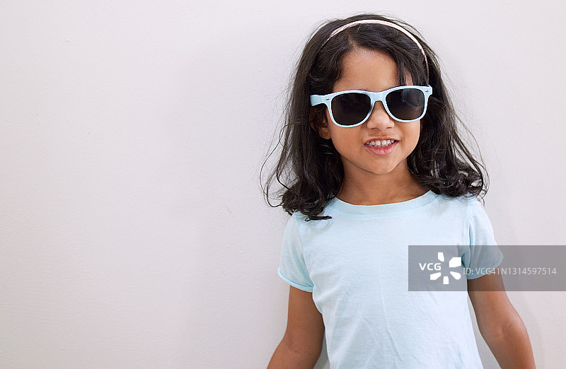 一个可爱的小女孩戴着太阳镜，靠墙站着图片素材