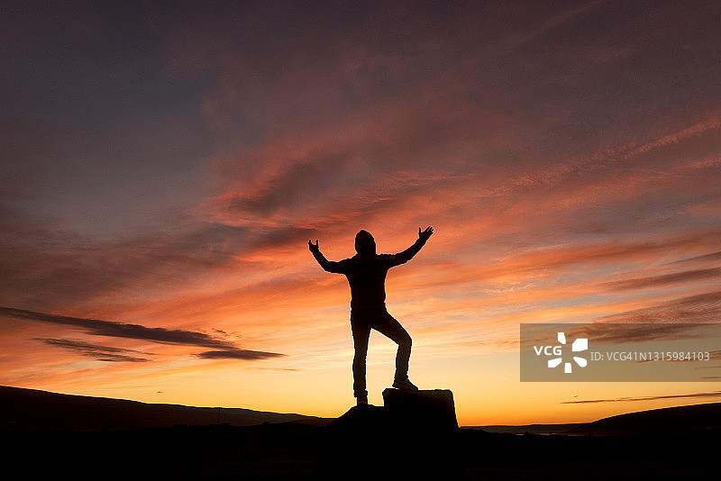 风景的剪影，一个快乐的人站在背包和举起的手臂在山顶上的背景多云的天空在彩色日落在夏季。旅游、攀岩、徒步旅行。图片素材