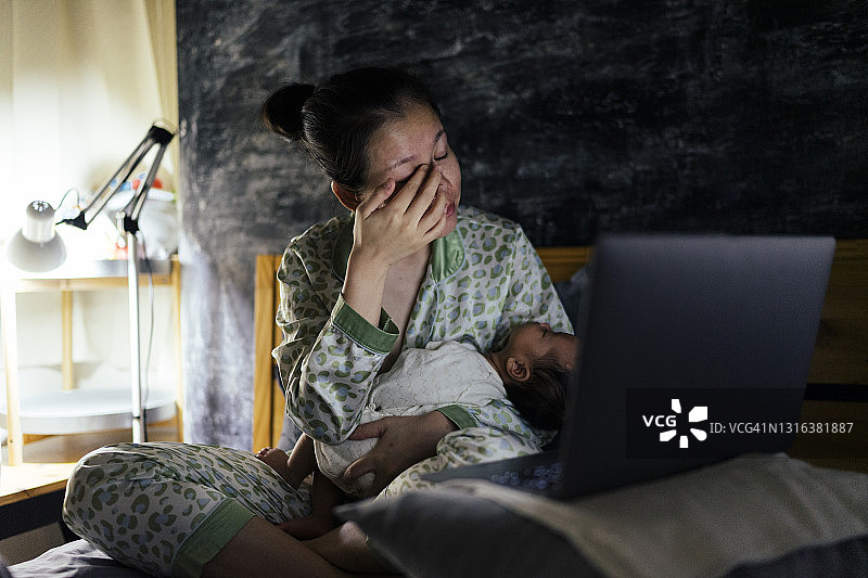 一位亚洲母亲深夜醒来给婴儿喂奶，然后在床上点击笔记本电脑图片素材