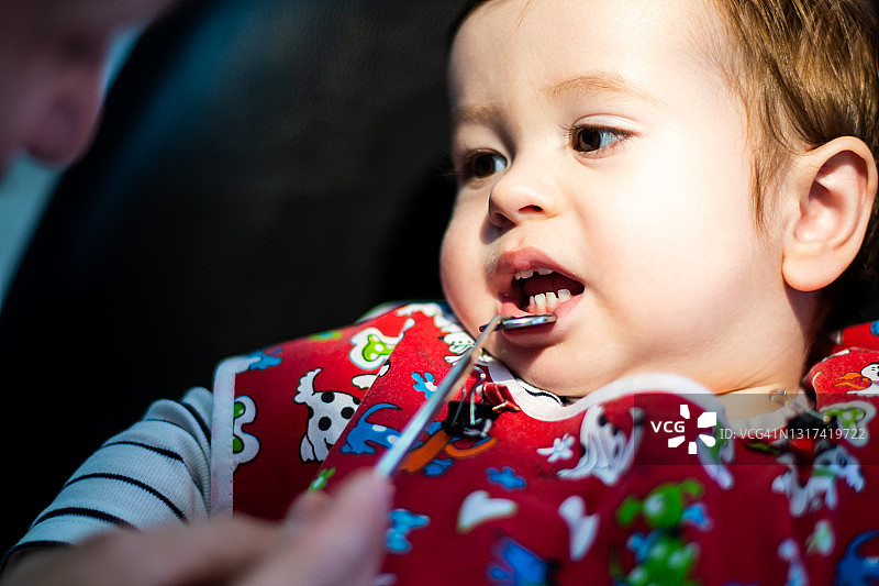 在牙医的办公室里，一个两岁以下的孩子穿着一件红色的长袍，看着牙医，牙医用一面小镜子照下了他的下嘴唇。选择性方式。图片素材