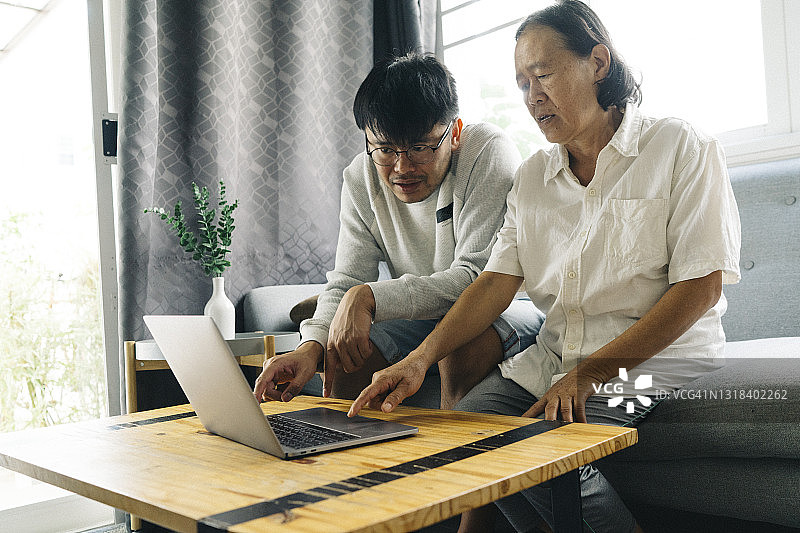 亚洲年轻的成年人儿子教年长的母亲如何使用社交媒体的笔记本电脑在家里图片素材