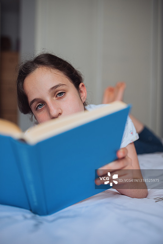 一个蓝眼睛的白人女孩躺在她的床上读一本非常有趣的书的特写。图片素材