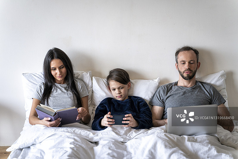 当父亲和儿子在床上使用无线技术时，母亲正在看书图片素材